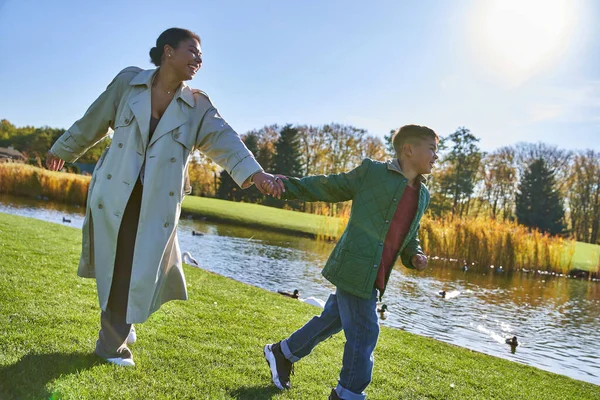 Feliz infancia, mujer afroamericana corriendo con su hijo cerca del estanque, trajes otoñales, temporada de otoño - foto de stock