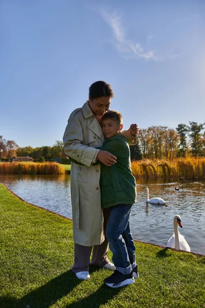 Amour d'enfant, heureux garçon étreignant mère près du lac avec des cygnes, famille afro-américaine, nature automnale — Photo de stock