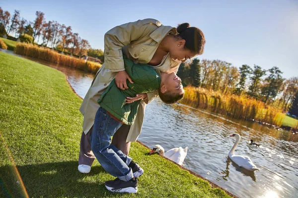 Amor familiar, madre afroamericana feliz y el niño se divierten, abrazos cerca del lago, la naturaleza, el otoño - foto de stock