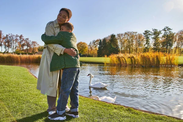 Amor infantil, niño feliz abrazando a la madre cerca del lago con cisnes, familia afroamericana, temporada de otoño - foto de stock