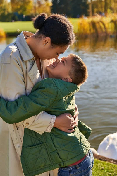 Amor maternal, niño alegre abrazando a la madre cerca del lago, familia afroamericana, temporada de otoño, otoño - foto de stock