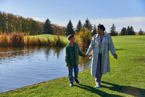 Мать и сын держатся за руки и ходят по траве возле озера, африканская американская семья, спокойный — стоковое фото