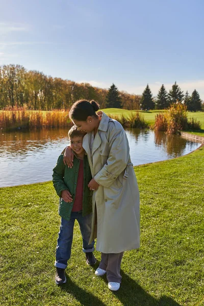 Счастливая мать обнимает сына, стоя возле озера в парке, африканская американская семья, осень моды — стоковое фото