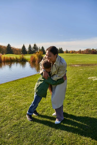 Happy woman hugging son, standing near lake in park, african american family, autumn fashion — Stock Photo
