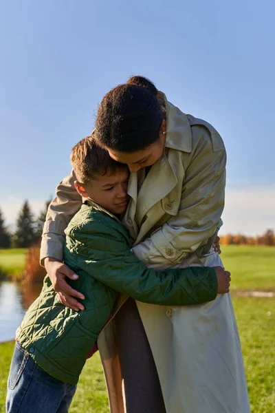 Donna afroamericana che abbraccia figlio, calmante, in piedi vicino al lago nel parco, famiglia afroamericana — Foto stock