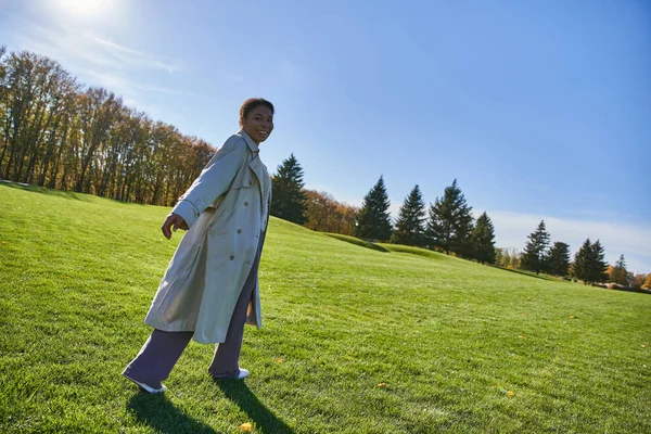 Heureuse femme multiraciale en trench coat marchant sur l'herbe dans le parc, mode automnale, journée ensoleillée — Photo de stock