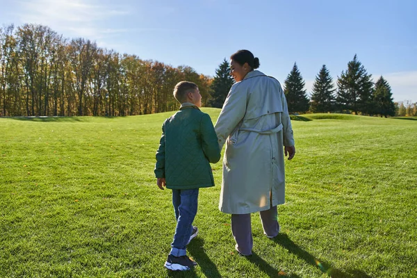 Vue arrière de la femme afro-américaine marchant avec son fils dans le parc, herbe verte, tenues d'automne, vêtements de dessus — Photo de stock