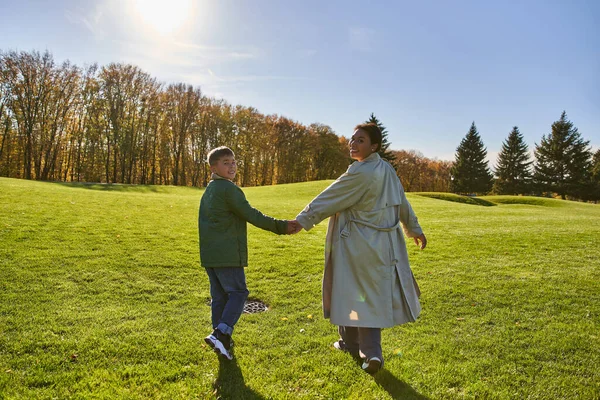 Día soleado, mujer afroamericana caminando con hijo en el parque, hierba verde, trajes de otoño, ropa de abrigo - foto de stock