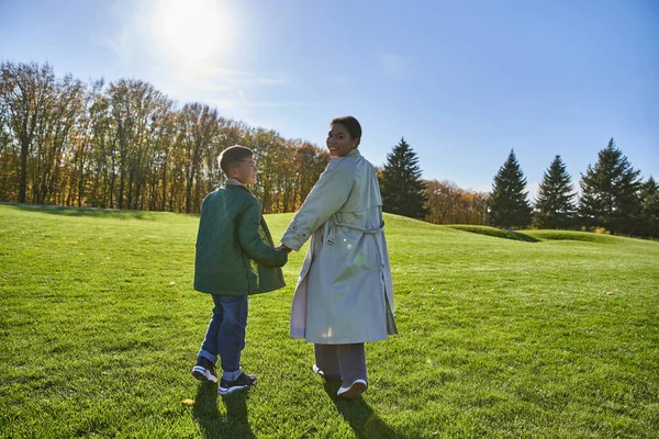 Sunny day in fall, african american woman walking with son in park, green grass, autumn, outerwear — Stock Photo