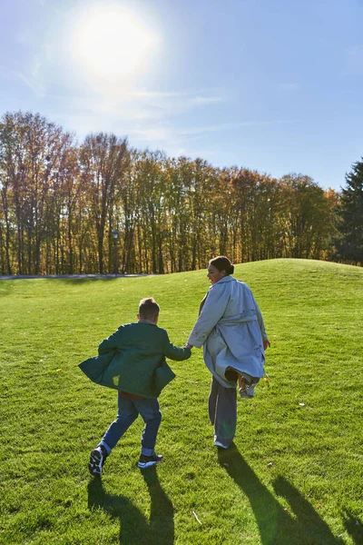 Journée ensoleillée à l'automne, femme afro-américaine courant avec son fils dans le parc, herbe, mode automnale — Photo de stock