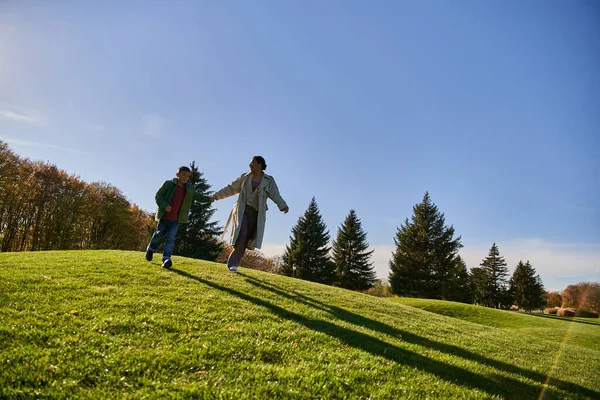 Sunny day in autumn, happy african american woman running together with son in park, candid — Stock Photo
