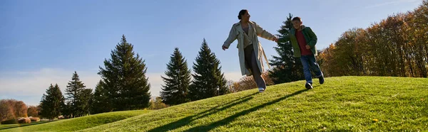 Día soleado en otoño, mujer afroamericana feliz corriendo junto con su hijo en el parque, cándido, pancarta - foto de stock