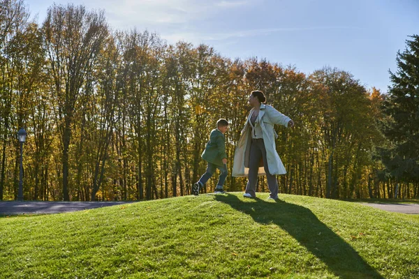 Радісна мати і син, афроамериканка, що стоїть на зеленому пагорбі з хлопчиком, сонячний день, осінь — стокове фото