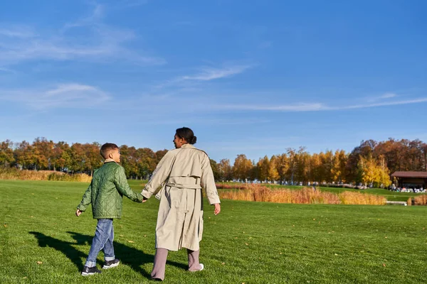 Joyeuse mère afro-américaine et son enfant se tenant la main, marchant sur l'herbe verte, chaudes journées d'automne — Photo de stock