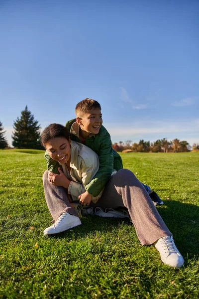 Feliz madre e hijo en el parque, día soleado, otoño, juguetón chico afroamericano abrazando a mamá, diversidad - foto de stock