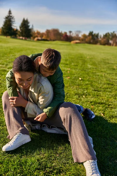 Feliz madre e hijo sentado en la hierba, día soleado, otoño, juguetón africano americano niño abrazando mamá - foto de stock