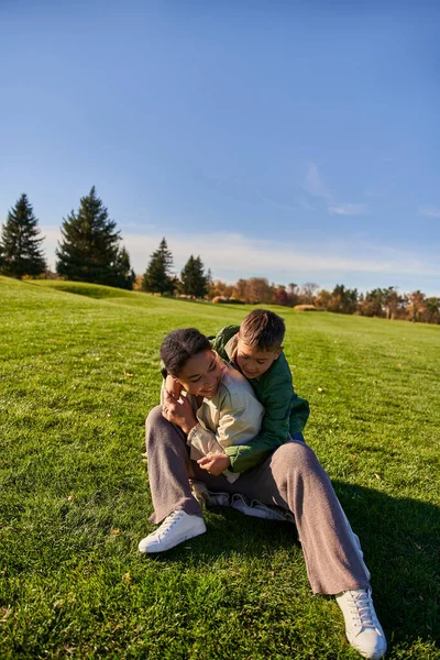 Joyful mother and son sitting on grass, sunny day, autumn, playful african american boy hugging mom — Stock Photo