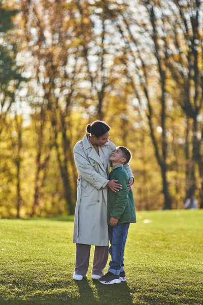 Alegre afroamericano madre e hijo abrazándose en el parque otoñal, mujer y niño en ropa de abrigo - foto de stock