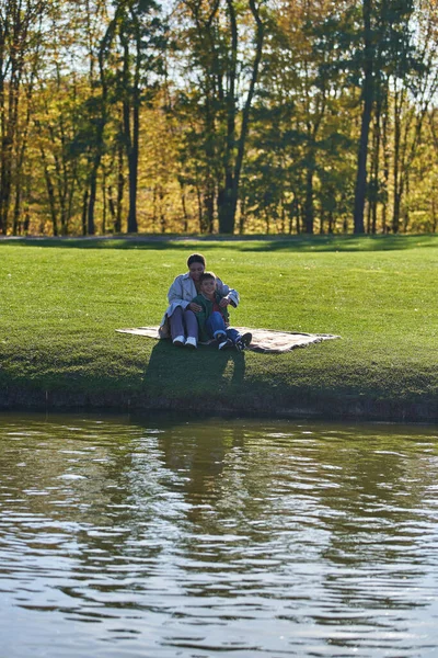 Joyeux mère et fils étreignant dans le parc automnal, assis sur la couverture près du lac, afro-américain — Photo de stock