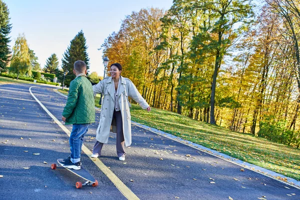 Heureux mère et fils, femme regardant garçon sur penny board, feuilles d'automne, famille afro-américaine — Photo de stock