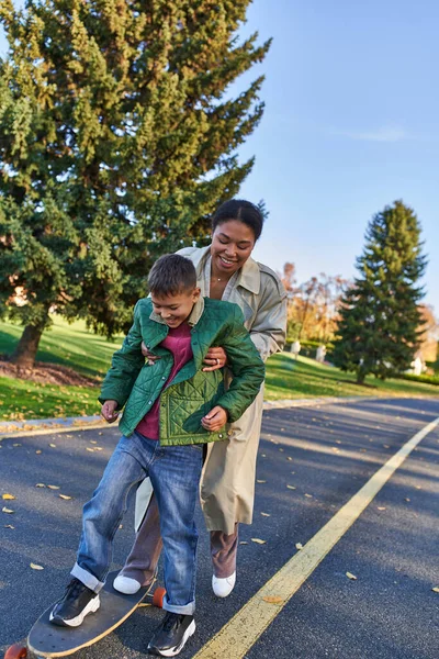 Madre e hijo en el parque de otoño, mujer afroamericana que apoya al niño en el tablero del centavo, momentos felices - foto de stock