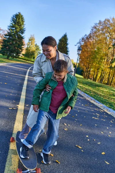 Mãe e filho no parque de outono, mulher afro-americana apoiando menino no penny board, momentos alegres — Fotografia de Stock