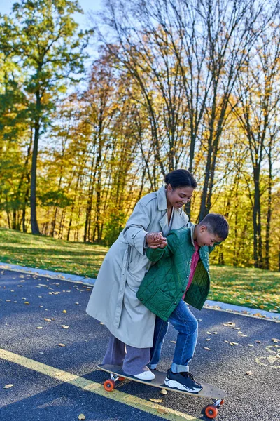 Mãe e filho, tempo de colagem, parque de outono, mulher americana africana andando perto do menino no penny board — Fotografia de Stock