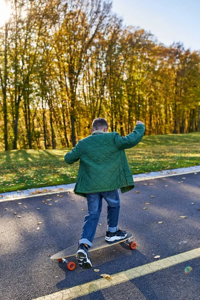 Visão traseira do menino em outerwear e jeans montando penny board no parque, outono, folhas douradas, garoto bonito — Fotografia de Stock