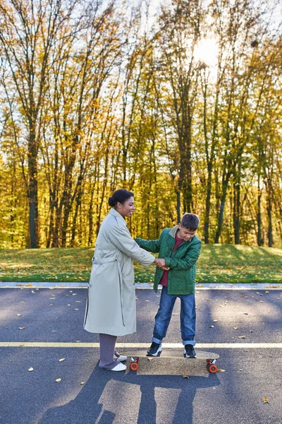 Mother and son in park, happy african american woman holding hands with boy on penny board, autumn — Stock Photo
