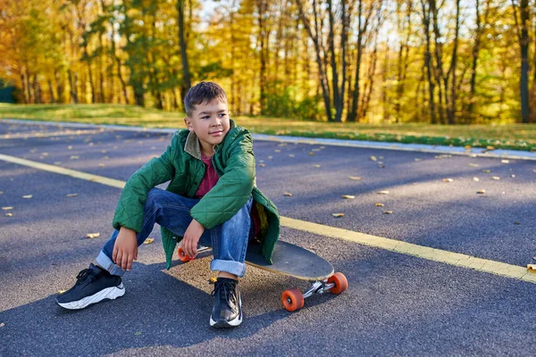 Pensivo menino americano africano em outerwear sentado em penny board, parque de outono, temporada de outono — Fotografia de Stock