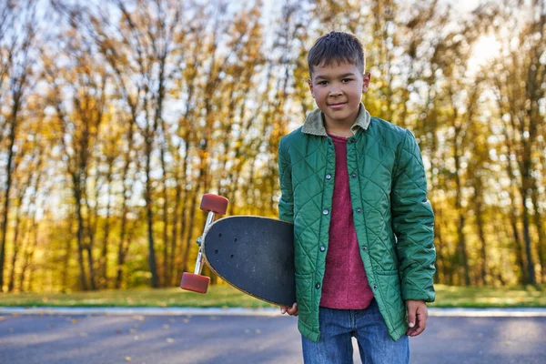 Menino americano africano feliz em outerwear segurando penny board, parque de outono, estação de outono, retrato — Fotografia de Stock
