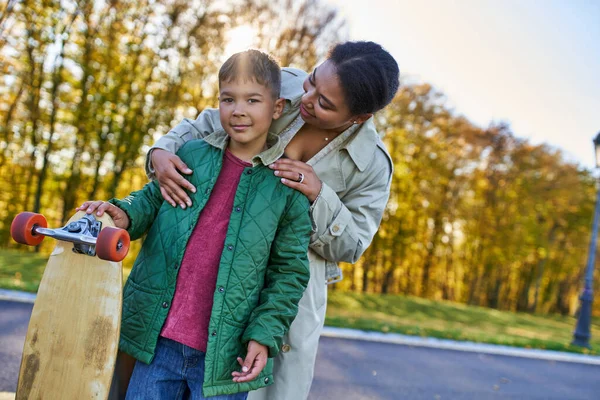 African american woman hugging son with penny board, autumn, fall season, motherly love, park — Stock Photo