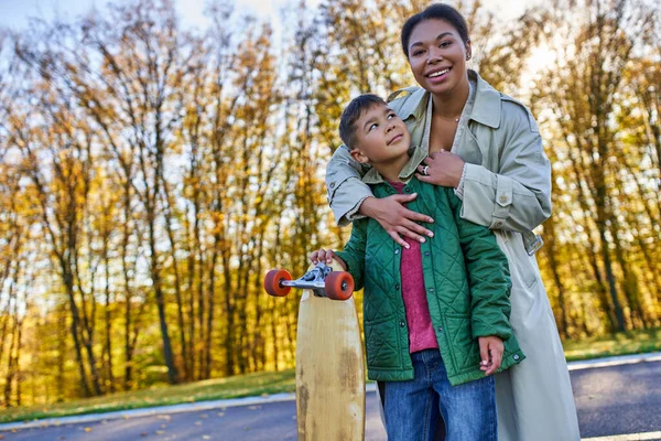 Heureuse femme afro-américaine étreignant son fils avec un penny board, automne, saison d'automne, amour maternel, parc — Photo de stock