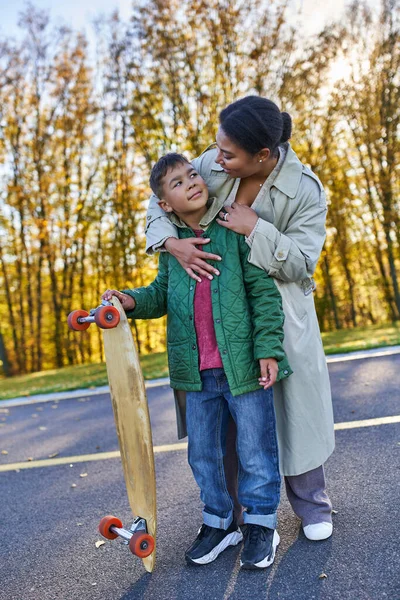 African american mother hugging son with penny board, autumn, fall season, motherly love, smile — Stock Photo
