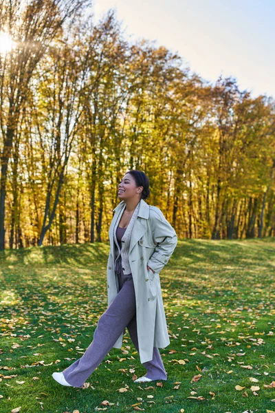 Mujer afroamericana feliz en gabardina caminando sobre hierba con hojas caídas, otoño, moda - foto de stock