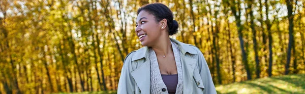 Cheerful african american woman in trench coat walking in autumn park, fashion outfit, banner — Stock Photo