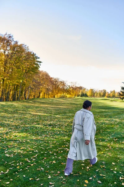 Vue arrière de la femme afro-américaine en trench coat marchant sur l'herbe aux feuilles dorées, parc d'automne — Photo de stock