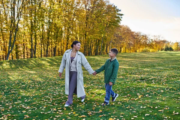 Mujer afroamericana feliz cogida de la mano con el hijo, caminando sobre la hierba con hojas doradas, otoño - foto de stock
