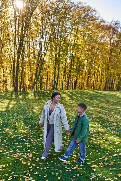 Fröhliche afrikanisch-amerikanische Frau hält Händchen mit Sohn, geht auf Gras mit goldenen Blättern, Herbst — Stockfoto