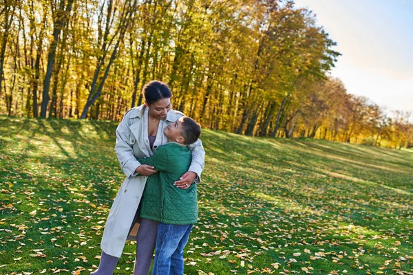 Mujer afroamericana feliz abrazándose con hijo, de pie sobre hierba con hojas doradas, otoño - foto de stock