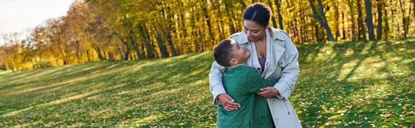 Mujer afroamericana feliz abrazándose con su hijo, de pie sobre hierba con hojas doradas, otoño, pancarta — Stock Photo