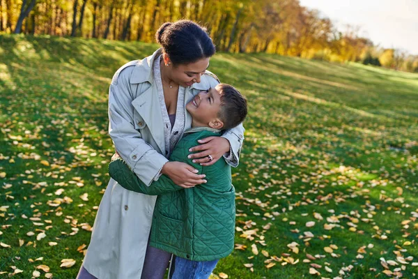 Mujer afroamericana feliz abrazándose con su hijo, de pie cerca de hojas doradas, el otoño, el amor maternal - foto de stock