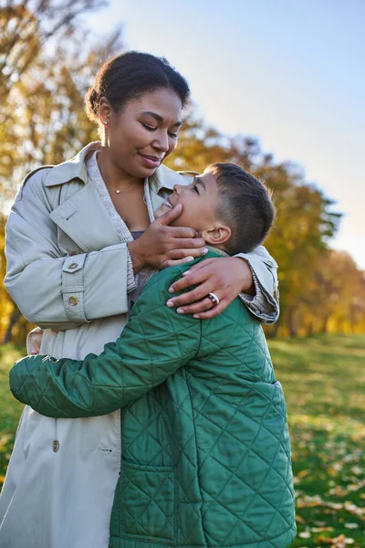 Amor maternal, alegría, mujer afroamericana abrazándose con su hijo, de pie cerca de hojas doradas, otoño - foto de stock