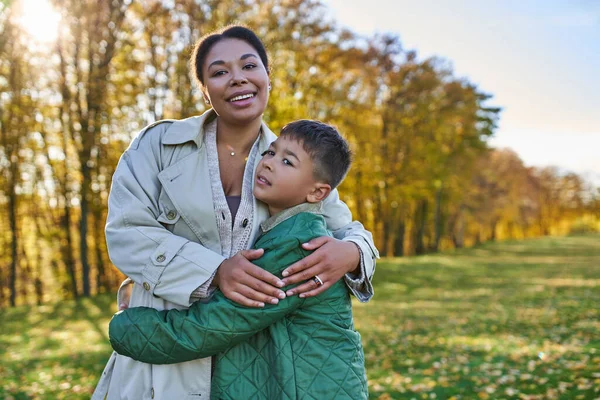 Motherly love, joy, african american woman hugging cute son, standing near golden leaves, autumn — Stock Photo