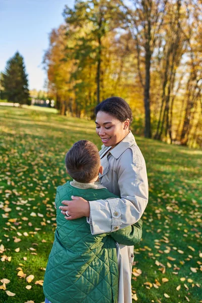 Amor maternal, mujer afroamericana feliz mirando a lindo hijo, de pie cerca de hojas doradas, otoño - foto de stock