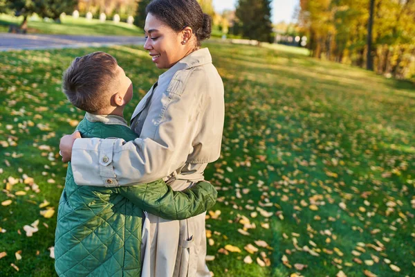 Motherly love, cheerful african american woman looking at son, standing near golden leaves, autumn — Stock Photo