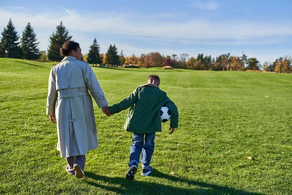 Chico afroamericano sosteniendo el fútbol, caminando con mamá en el campo verde, tomados de la mano, otoño - foto de stock