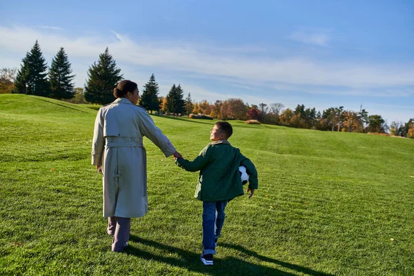 Chico afroamericano sosteniendo el fútbol, caminando con mamá en el campo verde, tomados de la mano, temporada de otoño - foto de stock