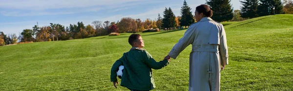 Chico afroamericano sosteniendo el fútbol, caminando con mamá en el campo verde, tomados de la mano, caída, bandera - foto de stock
