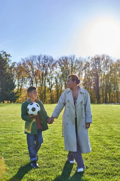 Chico afroamericano sosteniendo pelota de fútbol, caminando con mamá en el campo verde, tomados de la mano, otoño - foto de stock